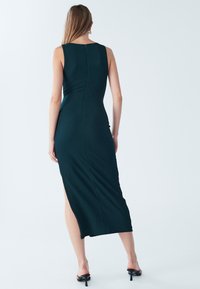 Dark green, ribbed, sleeveless midi dress with a side slit, featuring a high neckline and a concealed back zipper. Smooth texture, fitted design.