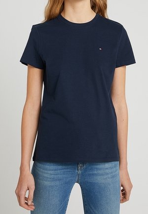 Kobieta ubrana w gładki granatowy t-shirt z krótkim rękawem i okrągłym dekoltem, w zestawieniu z jasnoniebieskimi jeansami, stoi na neutralnym tle.