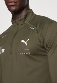 Donker olijfgroene sportieve half-zip top met wit logo, HYROX-tekst en opvallende stikdetails. Strakke pasvorm met een zachte textuur.