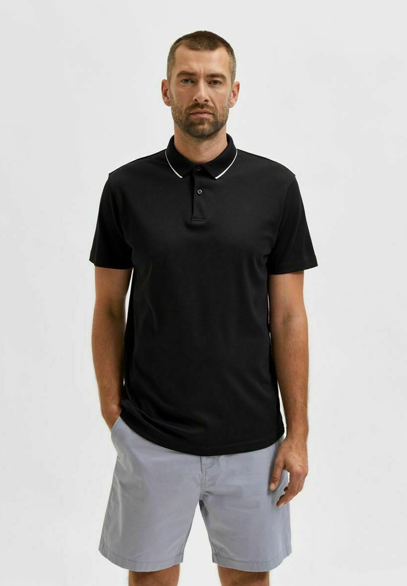 Polo negro de manga corta, con acentos en el cuello blanco y tapeta de tres botones; combinado con pantalones cortos gris claro. Textura suave.