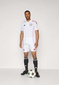 Hvid fodboldtrøje med polokrave, røde side-striber og logo på trøjen. Sorte shorts og sokker, med sorte støvler og en mønstret bold.