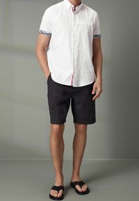 Camicia bianca a maniche corte con bordo blu a motivo ai polsini, abbinata a pantaloni corti neri e infradito nere, in piedi contro uno sfondo grigio.