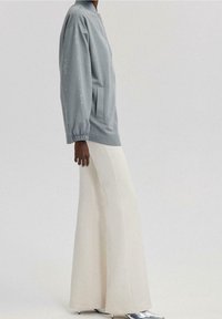 Lys grå bomberjakke med elastiske manchetter, prydet med diskrete broderede mønstre, parret med højtaljede, vide hvide bukser og metalliske sko.