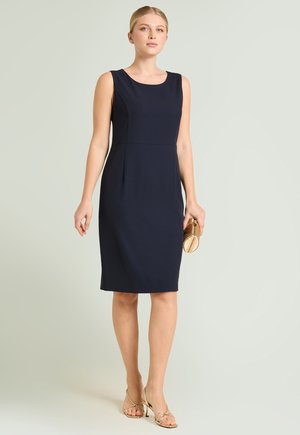 Vrouw draagt een mouwloze marineblauwe jurk tot op de knie, houdt een gouden clutch vast, met gouden sandalen met bandjes tegen een neutrale achtergrond.