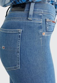 Jeans de denim azul con un diseño ajustado, que presentan un bolsillo trasero con un pequeño logotipo bordado y detalles de remaches en tono cobre.