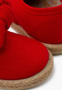 Sarkani kanvasu espadrilles ar noapaļotu purngalu, teksturētu virsmu un izpītas zāles detaļām uz zoles. Sānā ir redzams mezglota akcenta elements.