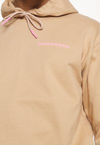 Felpa con cappuccio color camoscio realizzata in tessuto morbido, con coulisse rosa e logo "SCOTCH & SODA" in rosa sul petto.