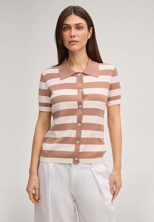 Femme portant un polo à manches courtes, rayures horizontales marron et blanches, boutonné, associé à un pantalon blanc taille haute, debout devant un fond uni.