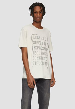 Lichtgrijze t-shirt met een grafische print met tekst in donkergrijs. Het shirt heeft een ronde hals en korte mouwen. Gecombineerd met donkere jeans.
