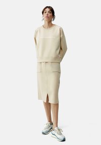 Beige sweatshirt with crew neck, featuring a subtle logo. Matched with a fitted beige skirt, knee-length, with horizontal pockets and a slit.