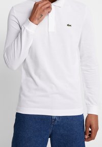 Vit långärmad polo skjorta i texturerad bomull, med klassisk krage, treknappsstängning och en liten grön logotyp på bröstet.