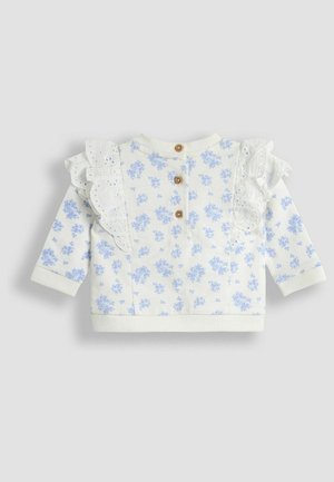 Weißes Langarmoberteil mit blauem Blumenmuster, ausgestattet mit Rüschendetails, drei Holzknöpfen im Rücken sowie gerippten Bündchen und Saum.