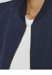 Marinblå bomberjacka med ribbad krage och guldfärgad dragkedja, med en slät textur. Vit skjorta syns under.