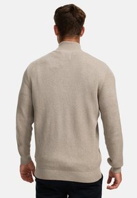 Lichtbeige trui met een textuur, gebreid patroon. Heeft een hoge kraag en ribgebreide boorden en zoom. Zichtbare naaddetails bij de schouders.
