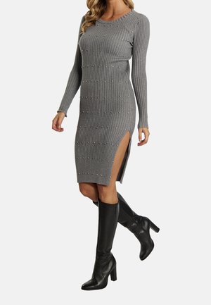 Femme portant une robe en maille côtelée grise avec des détails en perles et une fente latérale, associée à des bottes noires à talons montantes jusqu’aux genoux.