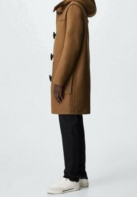 Cappotto di lana marrone con cappuccio, chiusure a toggles e tasche laterali. Indossato su pantaloni scuri e sneakers bianche. Silhouette semplice e dritta.
