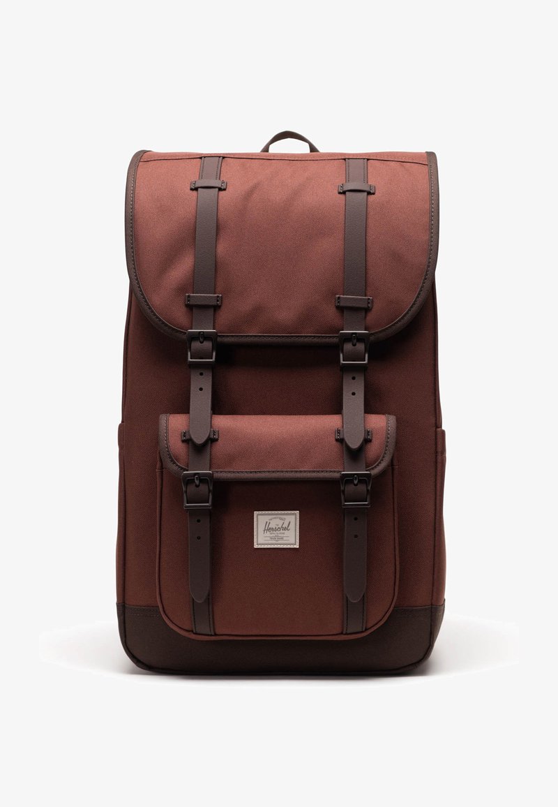 Sac à dos en tissu marron avec des sangles et des boucles en cuir foncé, poche avant et étiquette de marque Herschel sur fond blanc.