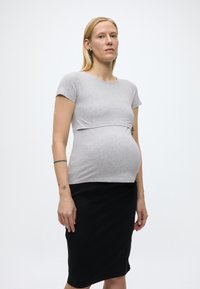 Těhotná žena s blond vlasy, oblečená v přiléhavém šedém tričku s krátkým rukávem a černé sukni po kolena, stojící před jednoduchým bílým pozadím.