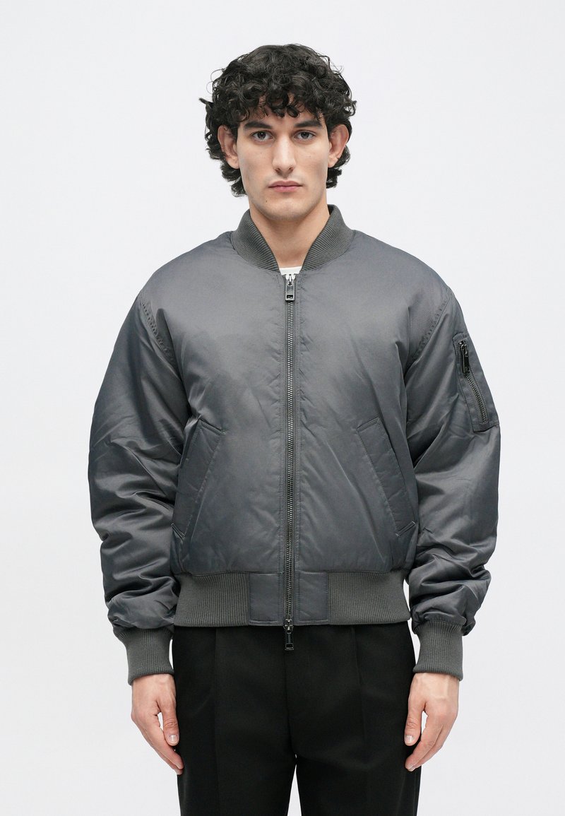 Grijze bomberjack van gladde stof, met een rits aan de voorkant, ribbelmanchetten en een ritszak op de linkermouw.