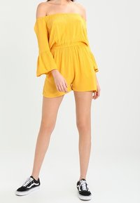 Gul off-shoulder rompers med trumpetärmar, rynkad midja och shorts. Matchad med svarta sneakers med vita snören och sulor.