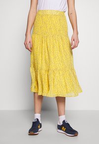 Person iført en gul blomsterkjole i midi-længde, hvide sokker og navyblå samt gule sneakers, der står mod en ensfarvet baggrund.