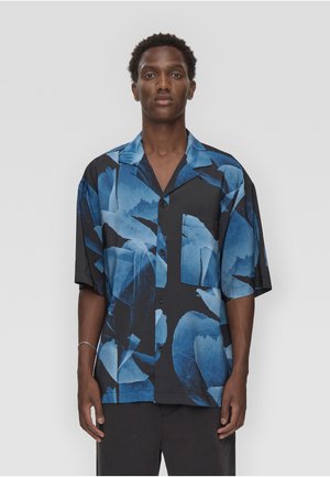 Mand iført en løs sort knap-op skjorte med store blå abstrakte blomsterprint, stående mod en ensartet lys grå baggrund.