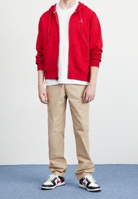 Sudadera roja con cremallera y cordones, llevada sobre una camiseta blanca. Pantalones beiges y zapatillas negras y blancas con detalles rojos completan el atuendo.