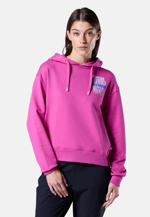 Vrouw met een helderroze hoodie met voorzak en zwarte broek, staand met ontspannen handen tegen een effen lichtgrijze achtergrond.
