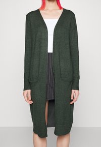 Mörkgrön stickad cardigan med öppen front, långa ärmar, två framfickor och avslappnad passform, kombinerad med en vit topp och randig kjol.