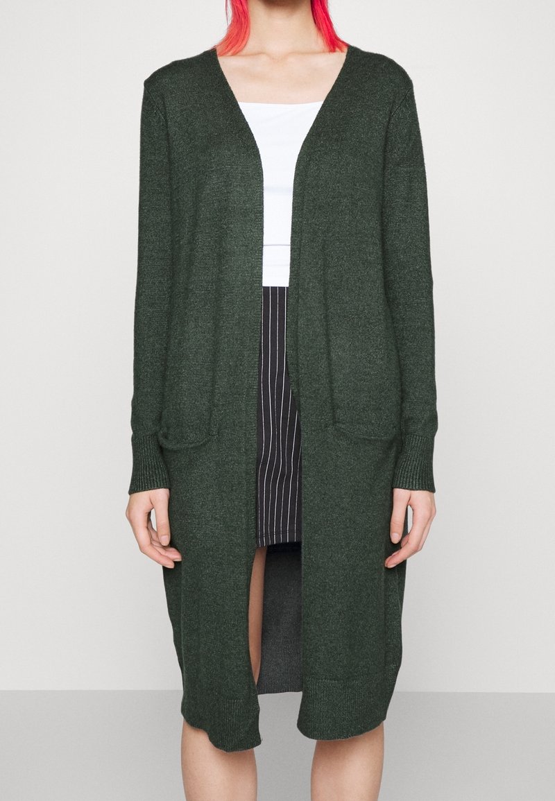 Mörkgrön stickad cardigan med öppen front, långa ärmar, två framfickor och avslappnad passform, kombinerad med en vit topp och randig kjol.