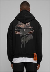 Sudadera negra corta con capucha y un estampado gráfico de un sombrero estilo pescador camuflado y texto "rostros olvidados" en la parte trasera. Cuenta con una etiqueta de tela naranja.