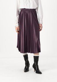 Gonna a pieghe viola in tessuto satinato, con vita alta e una consistenza liscia. Abbinata a stivali neri con tacco.