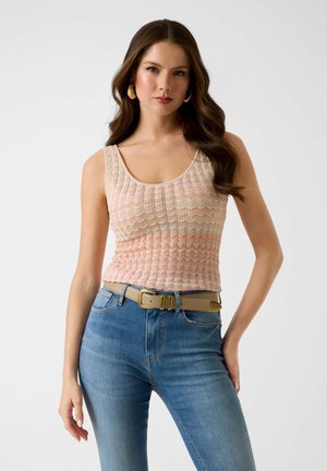 Frau mit langen braunen Haaren, die goldene Creolen, ein ärmelloses beiges Stricktop, einen hellbraunen Gürtel und blaue Jeans trägt und mit einer Hand auf der Hüfte posiert.