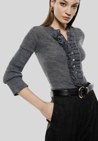 Maglione grigio lavorato a maglia con dettaglio arricciato sul davanti, chiusura con bottoni e maniche a tre quarti, abbinato a pantaloni neri a vita alta con cintura.