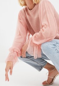 Ljust rosa stickad tröja med genomskinliga puffärmar och elastiska muddar, kombinerad med blå högmidjade jeans och remsandaler.