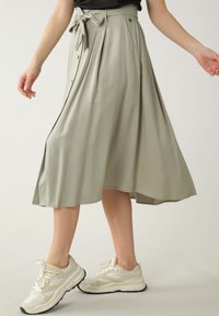 Jupe midi vert clair avec lien à la taille, détails de plis, texture lisse et boutons sur le côté. Assortie avec des baskets blanches et beiges.