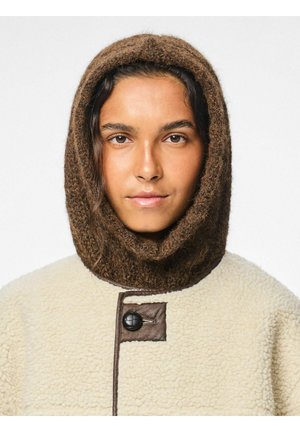 Giovane con cappuccio in maglia marrone e giacca in sherpa crema con rifiniture in pelle e grande bottone nero, volto in avanti su sfondo bianco.
