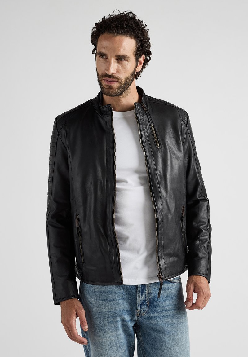 Zwarte leren jas met een hoge kraag, voorzien van ritszakken en gestructureerde schouderdetails. Draag het over een wit shirt.