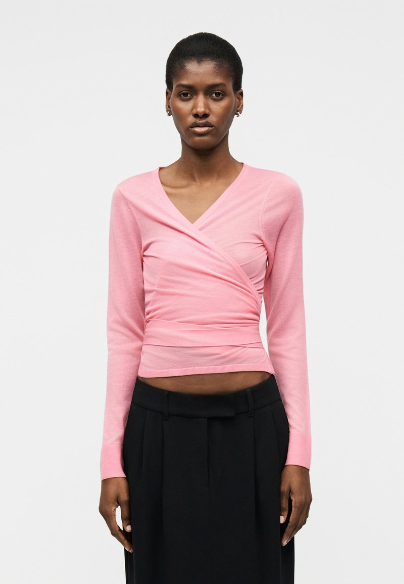 Femme portant un haut ajusté rose à manches longues de style cache-cœur et un pantalon noir taille haute, debout devant un fond clair uni.