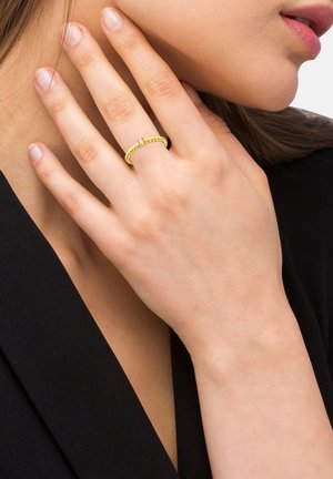 Bague en or avec un design délicat, comportant une rangée de petites pierres claires serties dans un bandeau texturé, portée sur une main contre un fond sombre.
