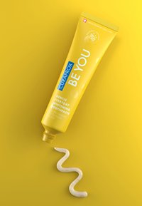 Tube de dentifrice jaune avec du texte bleu étiqueté "CURAPROX BE YOU." Un fil de dentifrice blanc avec des éclats orange s'étire le long d'un fond jaune.