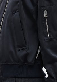 Κοντινό πλάνο μαύρου σακακιού που δείχνει μια ασημένια τσέπη με φερμουάρ στο μανίκι, μια πλαϊνή τσέπη, ριμπ ύφασμα στρίφωμα και μια μικρή εμφανή ετικέτα υφάσματος της μάρκας.