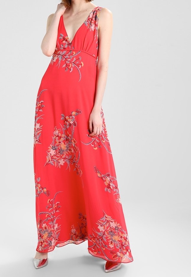 Femme portant une robe maxi rouge sans manches avec des motifs floraux et des talons pointus argentés, debout devant un fond uni gris clair.