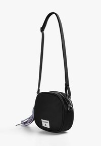 Bolso cruzado negro hecho de tela, de forma redonda, con cierre de cremallera, correa ajustable, que presenta acentos de borlas coloridas y un parche con logo.