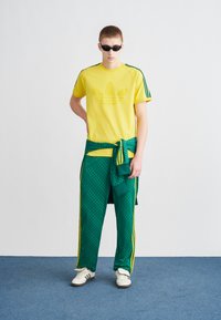 Gelbes T-Shirt mit strukturiertem Logo, grüne Trainingshose mit Muster und weiße Turnschuhe. Schwarze Sonnenbrille vervollständigt den sportlichen Look.