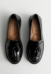 Mocasines de charol negro con punta redonda, detalle decorativo de lazo y plantilla acolchada en tono marrón. Acabado suave y brillante, diseño fácil de poner.