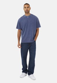 T-shirt blu oversize con scollatura rotonda e maniche corte, abbinata a jeans di jeans scuri e sneakers bianche, in piedi contro uno sfondo semplice.