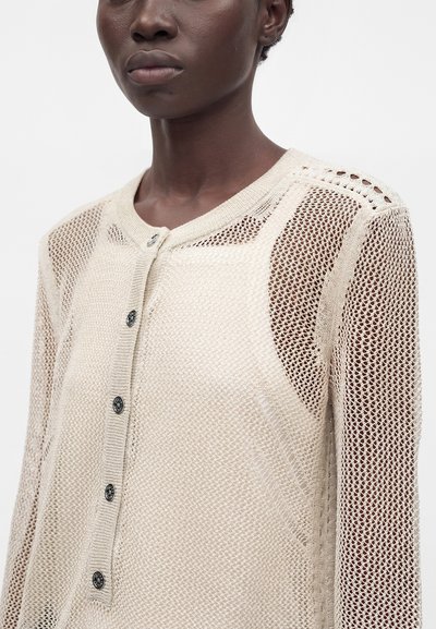 Gros plan d'une personne portant un cardigan en maille beige avec un devant boutonné et des manches en mesh.