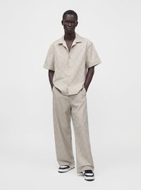 Homme portant une chemise beige à manches courtes boutonnée et un pantalon large assorti, avec des baskets noires et blanches, debout les mains dans les poches.