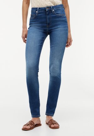 Kvinde iført jeans med mellemhøj talje i blå skinny-fit og brune flade sandaler med stropper, stående mod en enkel hvid baggrund.